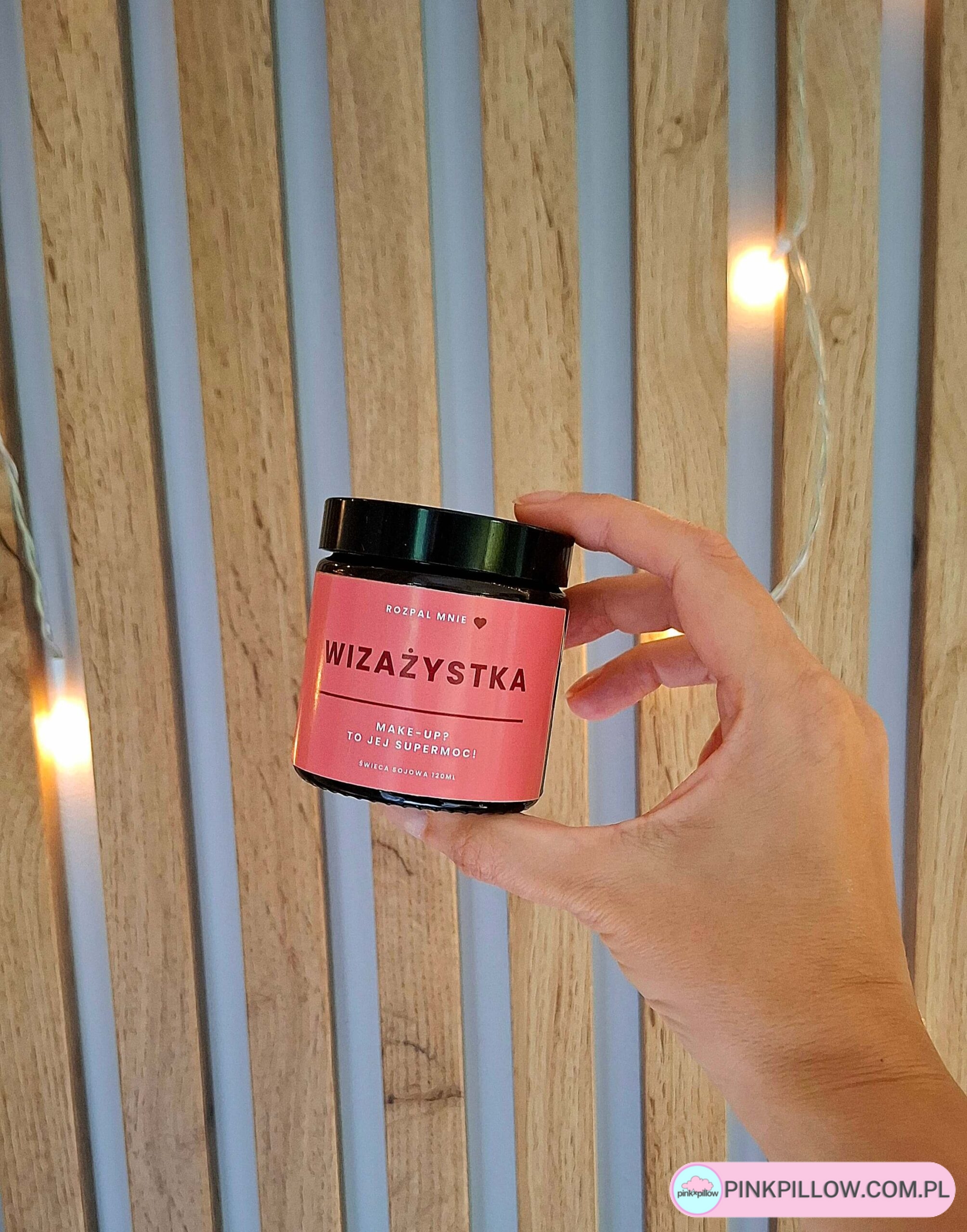 Kobieca dłoń trzyma świeczkę sojową dla wizażystki na tle drewnianych paneli i rozpalonych lampek.